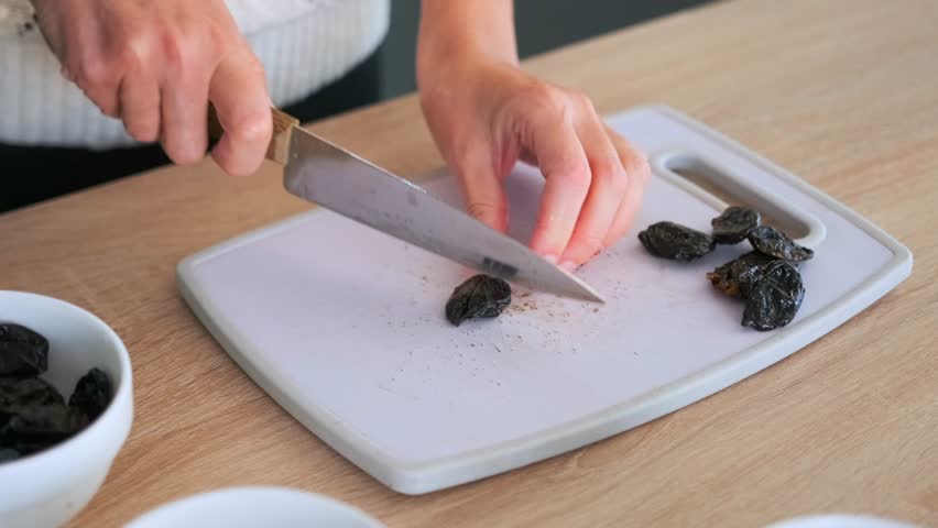 Close up of female hands cutting prunes with sharp knife on white cutting board on kitchen table. The process of preparing ingredients for a dish of porridge or smoothie.