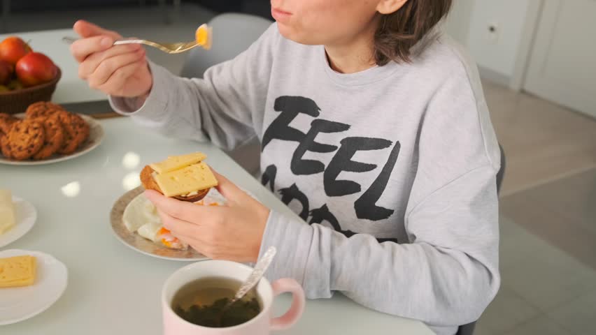 Happy young woman having breakfast in kitchen smiling at guy handing her sandwich with butter eating eggs with appetite sitting with coffee at table.
