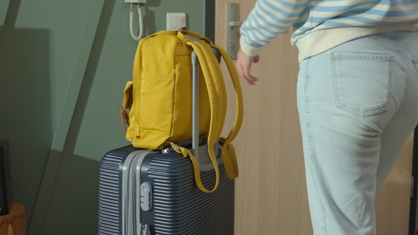 Woman puts on yellow backpack, taking suitcase and leaving apartment door. Person exit home with bag and luggage. Female traveler wearing jeans and sweater preparing for travel or departure indoors