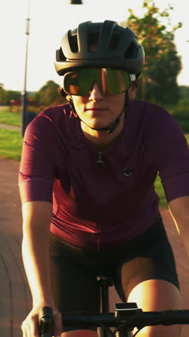 Portrait of young woman in black helmet and sunglasses is cycling on bicycle in summer park at sunset. Female is riding bicycle on cycle path. Vertical video