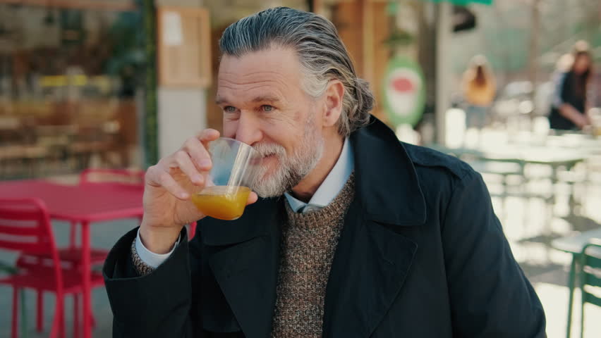 A mature man with a beard and long gray hair sits at an outdoor cafe, enjoying a glass of orange juice and looking around.