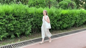 Young woman in a white dress walks along a city path next to a lush green hedge. She carries a beige tote bag. Summer fashion, urban lifestyle, and eco-friendly vibes. - Powered by Shutterstock - Get 15% off with code: PIKWIZARD15