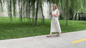 A woman in a linen dress walks along a sidewalk, using her smartphone. She strolls past weeping willows and a graffiti-covered wall. A tranquil summer scene. - Powered by Shutterstock - Get 15% off with code: PIKWIZARD15