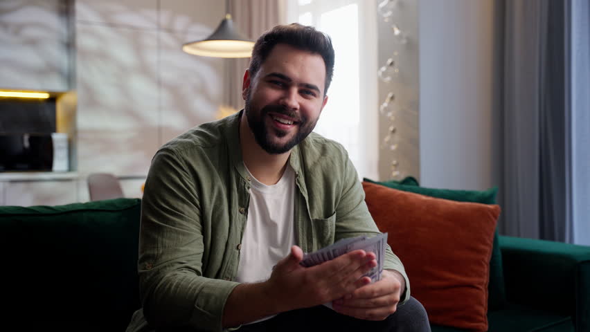 Happy smiling rich joyful Caucasian business man male guy homeowner client businessman customer sitting sofa winning lottery bet holding cash money dollars stack looking at camera smile at home wealth