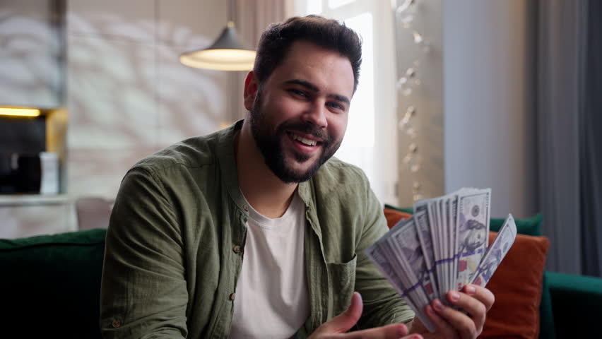 Happy smiling rich joyful Caucasian business man male guy homeowner client businessman customer sitting sofa winning lottery bet holding cash money dollars stack looking at camera smile at home wealth