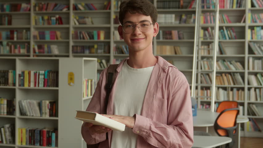 Teenage guy smiling happy looking at camera holding book buying literature prepare for reading in books store library in high school college studying learning Caucasian man male student education