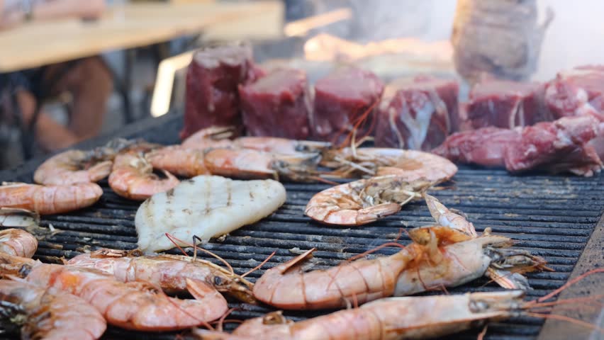 Red king prawns sizzle on a hot grill beside thick beef steaks. A cook uses metal tongs to flip the meat on the grate. Juicy seafood and beef grilling over open fire a delicious mix of surf and turf.