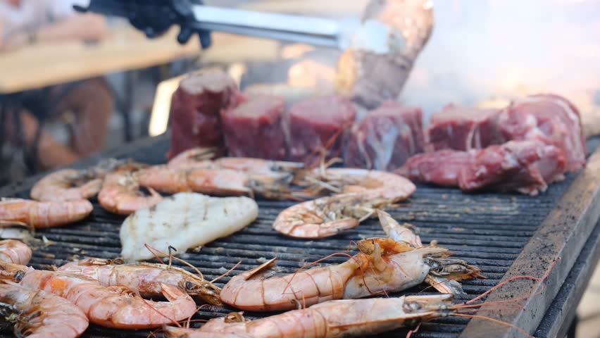 Red king prawns sizzle on a hot grill beside thick beef steaks. A cook uses metal tongs to flip the meat on the grate. Juicy seafood and beef grilling over open fire a delicious mix of surf and turf.