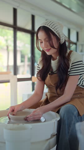 Young asian woman sculpting clay on spinning pottery wheel in ceramic workshop – relaxing creative hobby, art class activity, and handmade design practice in craft studio environment