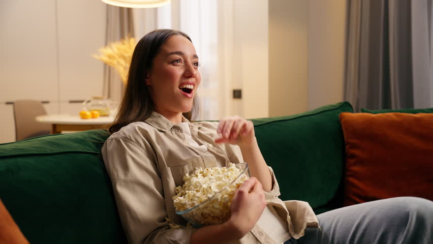 Caucasian woman girl falling on comfortable couch with bowl of popcorn turning on TV laughing enjoying snack watching television movie show cozy at home entertainment relaxing happy leisure weekend