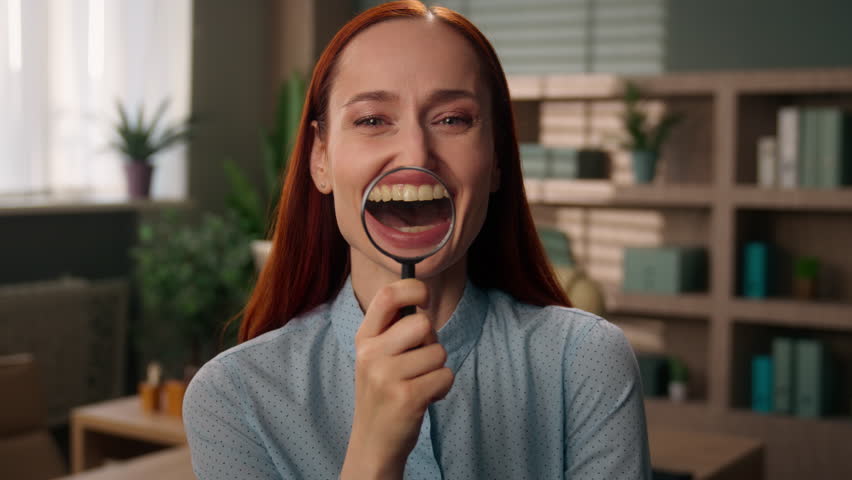 Caucasian business woman girl happy businesswoman holding magnifying glass magnifier near face investigator scientist research in office looking at camera smiling zooming teeth healthy smile laugh fun