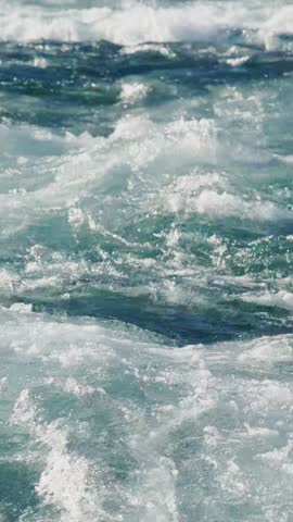 Turbulent waves crash against the rocky shoreline under clear blue skies, capturing the essence of a lively coastal landscape during daytime.