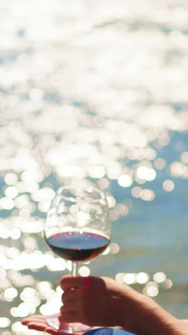 A hand holds a wine glass filled with red wine, shimmering in the light of the setting sun reflecting off the water. The atmosphere is relaxed and serene, perfect for unwinding.