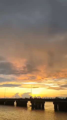 A beautiful sunset over the sea. The sky is orange and gray. There is a long pier with people standing on it. The water is calm and reflects the colors of the sky.