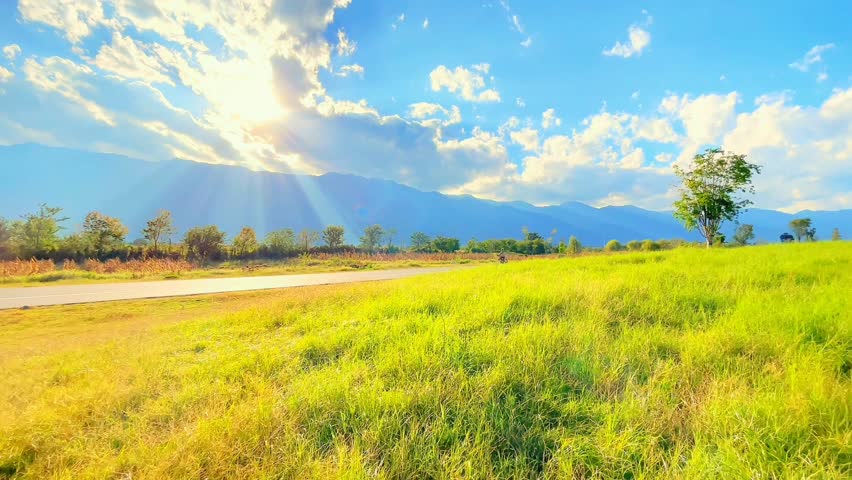 Peaceful countryside view with a person walking under a sunny blue sky, green fields, and distant mountains.

