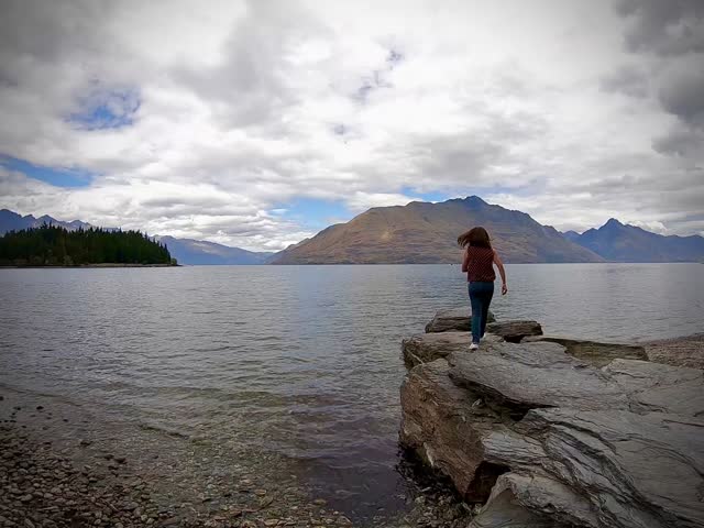 A lady walking on the rocks Overlooking Mountain and Lake Wakatipu Queenstown, Serene New Zealand Landscape with Rocky Shoreline and Cloudy Sky