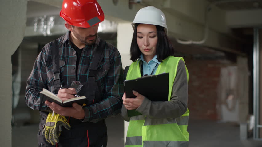 Multiethnic builders Japanese woman Caucasian man checking building write notes civil engineers in hardhat uniform check inspection clipboard talking industry business building project construction