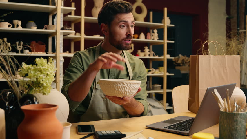 Happy joyful creative Caucasian talented craftsman man male guy businessman artisan small business owner holding pottery bowl decoration stoneware talking online video call laptop computer sell indoor