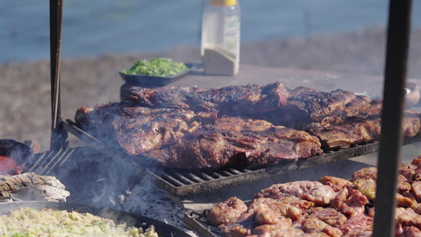 Still camera clip portraying Argentine asado type barbecue stake on fire, with ashes and smoke on the air and river in background.