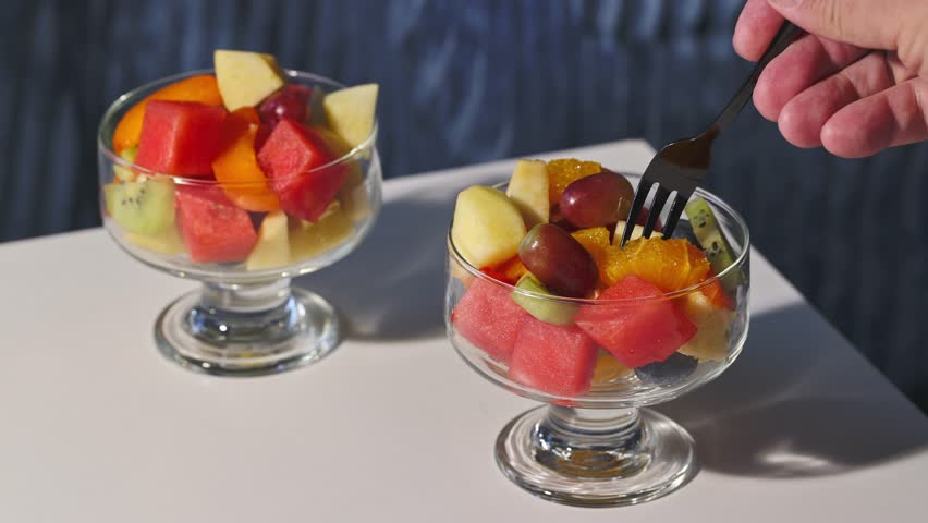 bowl of fresh fruit salad on blue background and hand with fork, food closeup, healthy eating