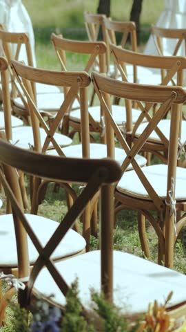 VERTICAL VIDEO Outside wedding ceremony in park among the trees. Dolly shot of wooden chairs with white pillows on it, slow motion. 