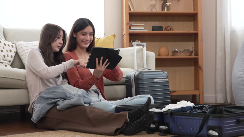 Two asian friends woman packing suitcase and choosing clothes together using tablet, planning a trip, checking fashion reviews or packing lists, travel preparation, content creator and travel blogger