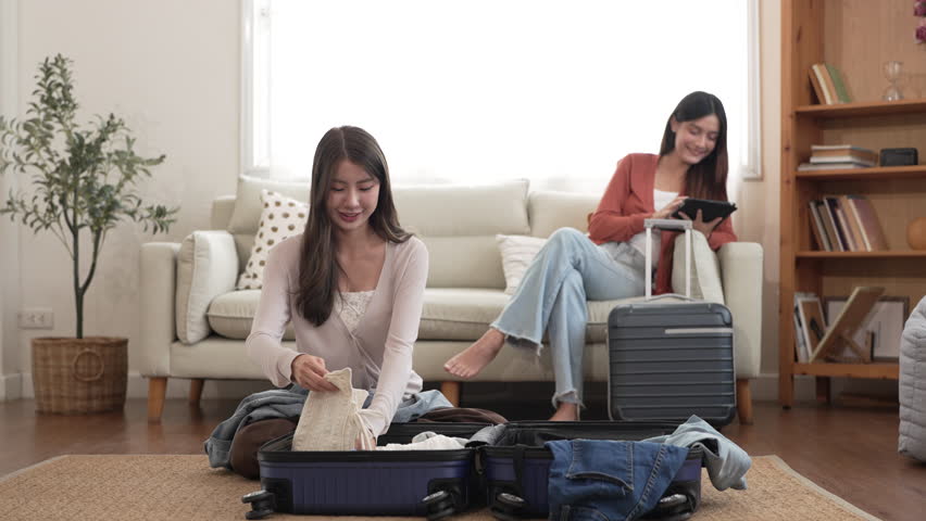 Young asian woman and friend preparing luggage packing clothes in suitcases for travel at home, planning holiday travel, content creator and travel blogger, friendship moments lifestyle vacation trip