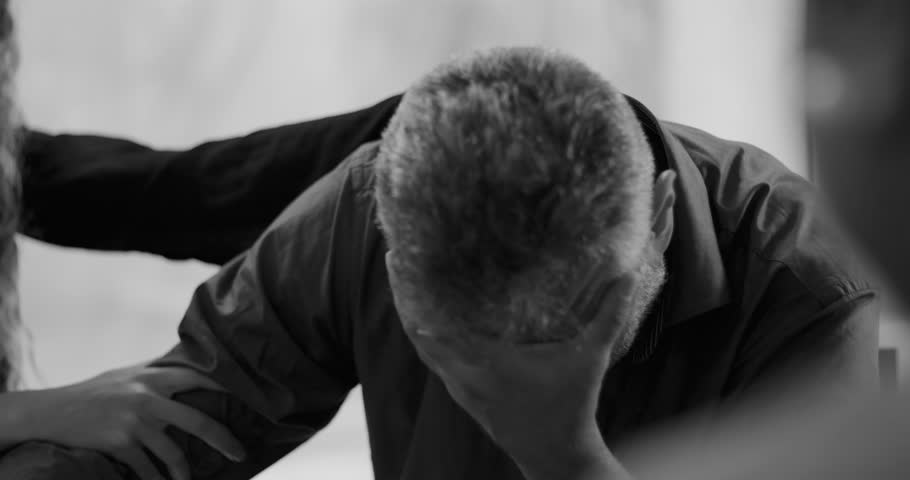 Man weeps during therapy session, covering face in hand while surrounded by supportive group members gently touching his arms in moment of deep emotional release