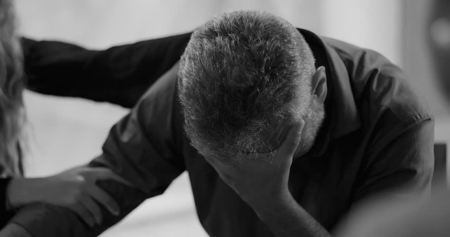 Man weeps during therapy session, covering face in hand while surrounded by supportive group members gently touching his arms in moment of deep emotional release