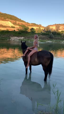 A beautiful, slender blonde woman in a swimsuit brought her horse for a swim at sunset.