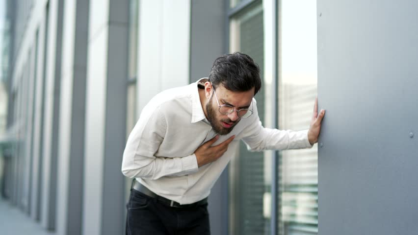 Businessman has heart attack, suffering from chest tension walking on street near office building. Upset unhealthy man holding sore spot with hands, breathing deeply, needs urgent medical attention