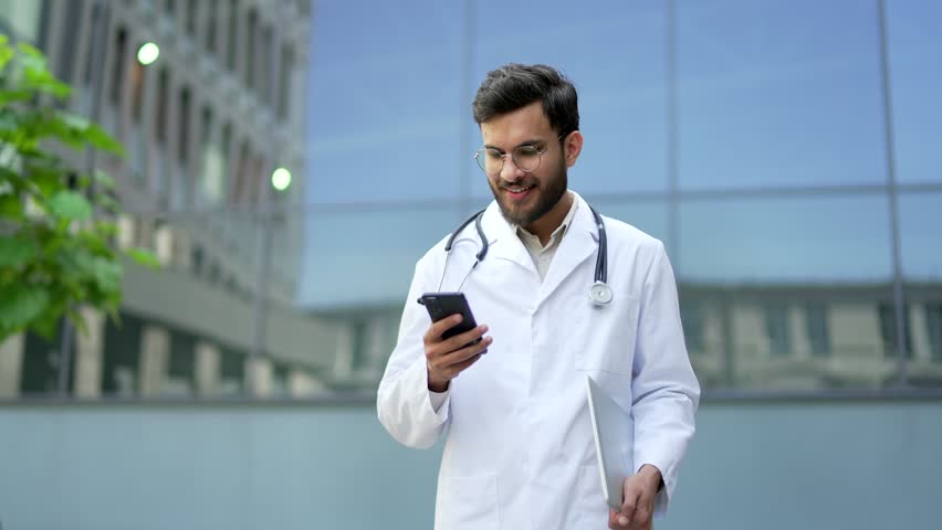 Smiling doctor in white coat using mobile phone walking outside near hospital building. Handsome medical worker physician checks smartphone, reads message, browsing web or chats online with patient