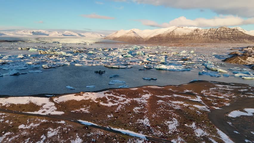 Drone shot showing icebergs floating on glacial lake surrounded by snowy mountains under blue sky in Iceland. Stunning view of frozen landscape and peaceful natural beauty in winter