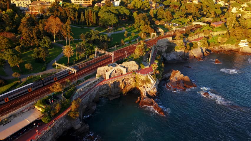 Golden hour drone shot of Genoa's coastline, showing lush parks, train tracks, and the sea