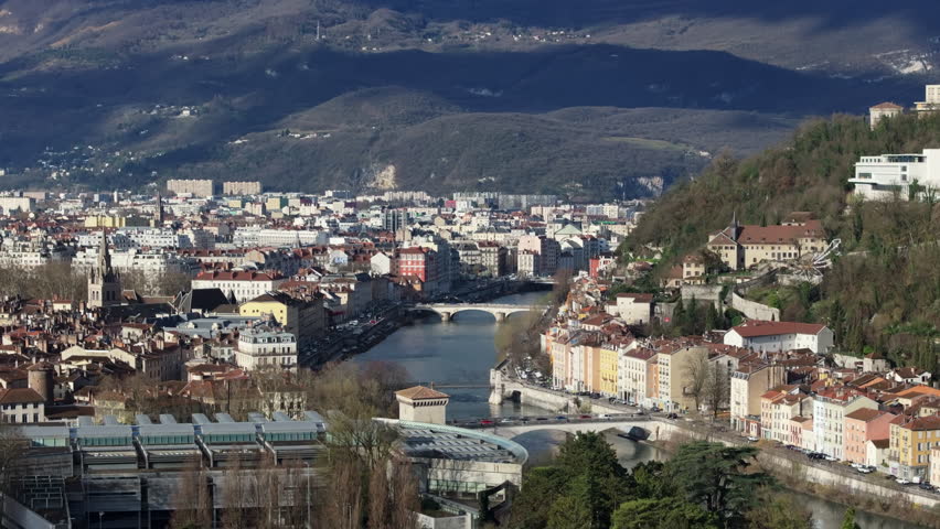 Aerial panorama of Grenoble city center, Isere river winding near Bastille hill, urban terrain framed by mountain ranges, revealing scenic cityscape