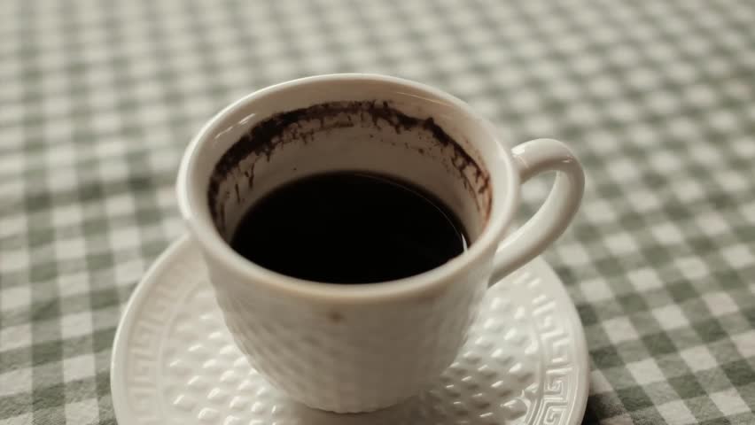 A close-up of a traditional Balkan coffee cup with grounds left inside, placed on a patterned saucer over a checkered tablecloth. Rustic and cultural morning ritual