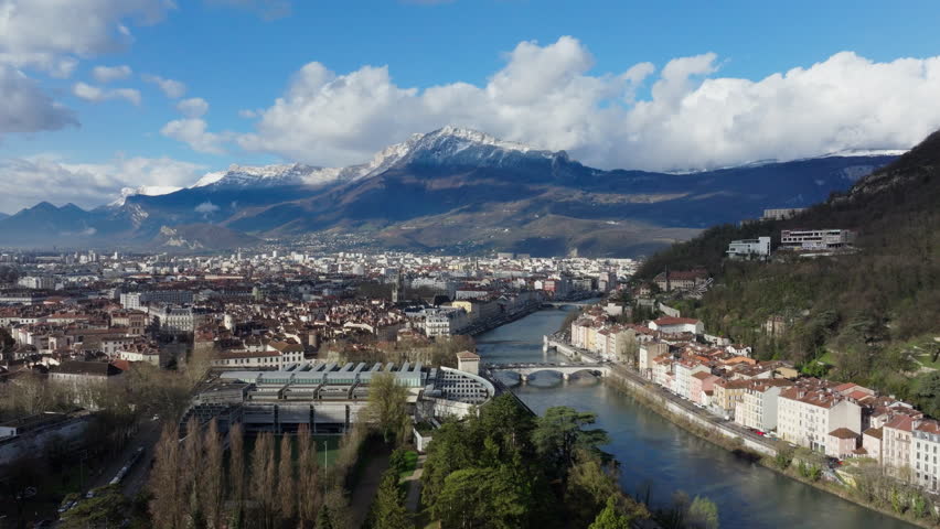 Panoramic aerial view of Grenoble city center, featuring bridges over Isere river and snowy mountains in the background