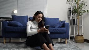 Young Asian businesswoman sitting on living room floor, experiencing emotional distress while reading devastating message on smartphone, displaying raw vulnerability and mental health struggles - Powered by Shutterstock - Get 15% off with code: PIKWIZARD15