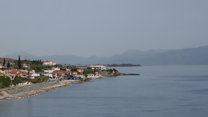Panoramic view of village in Greece showcasing beautiful coastal scenery with serene waters reflecting charming villages and mountains under bright sky. Pan camera moves to terracotta tiles rooftop