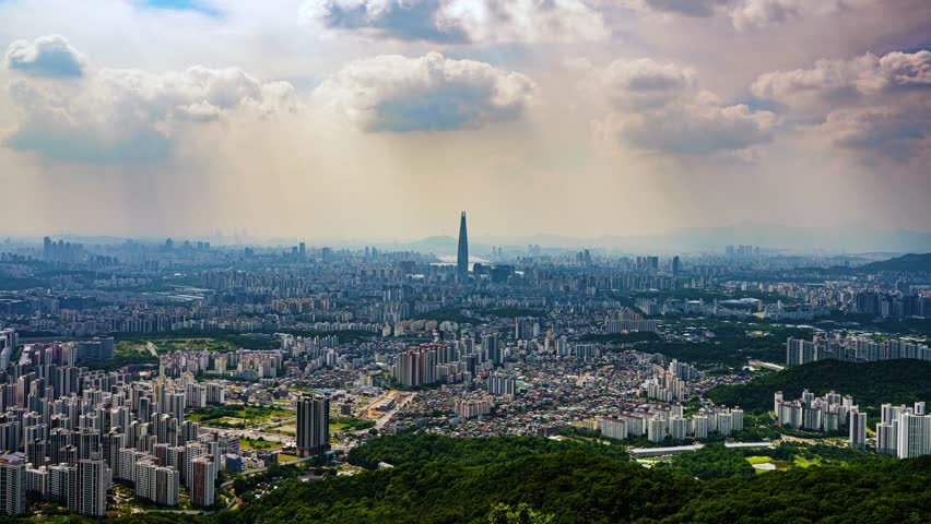 A dramatic timelapse shows clouds casting moving shadows over the vast urban sprawl of Seoul, South Korea, with the iconic Lotte World Tower standing tall in the center of the cityscape.
