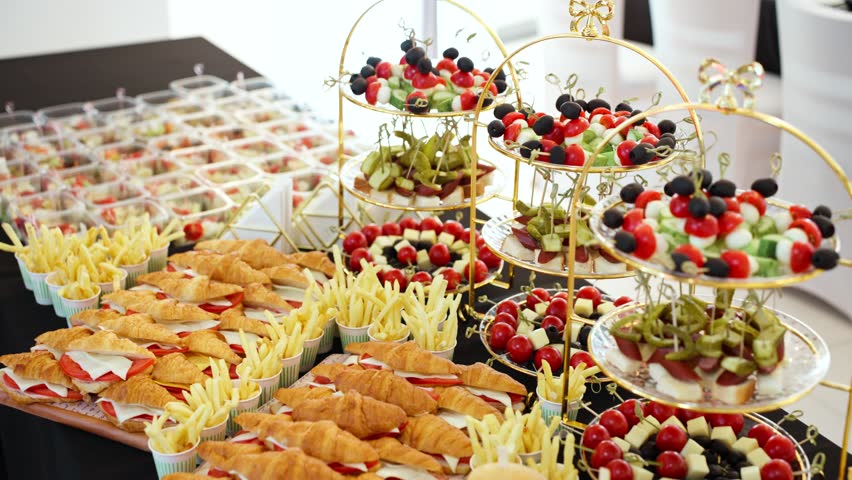 A slow dolly-in camera move pushes towards a delicious and varied catering buffet, showcasing croissant sandwiches, mini burgers, fries, and other appetizers for a special event.