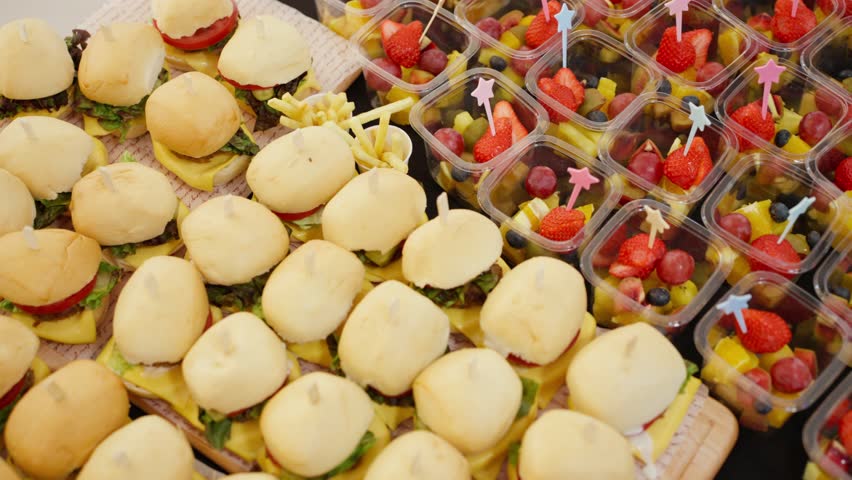 A beautifully arranged catering buffet table offers a delicious variety of fresh finger foods, including mini burgers, croissant sandwiches, fruit cups, and canapés for a party or event.