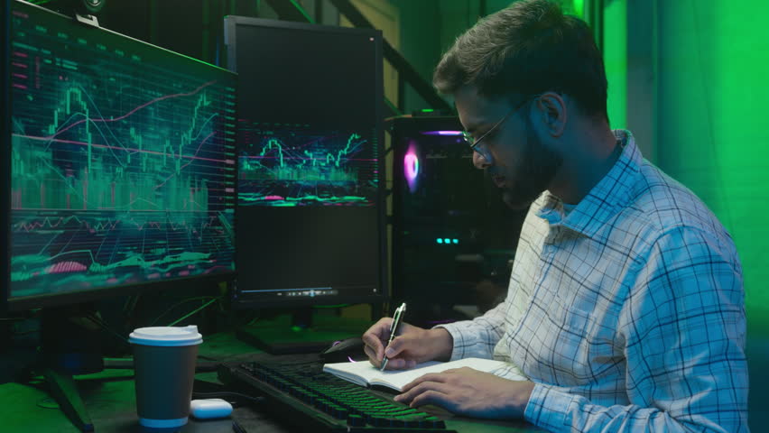 Indian Arabian man looking monitor computer chart graph trading online typing pc writing notes notebook analyst programmer male businessman business ethnic guy in neon office freelancer working crypto