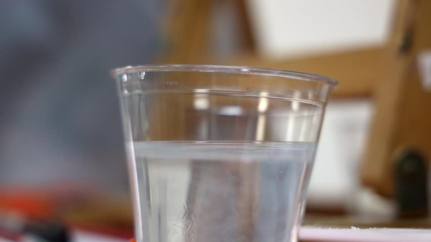 A closeup footage of cleaning paint brush in water in transparent cup, against blur background