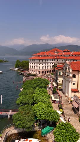 Drone video of tourists walking on Isola Bella, Lake Maggiore, Italy. Borromeo Palace with red-tiled roof shines under bright midday sun. Lush gardens and lake views complete this iconic destination