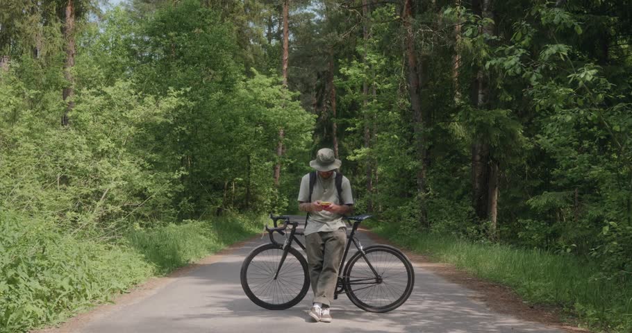 Millennial man in bucket hat stands with bicycle in the middle of a forest road, using his smartphone. Still camera shot. Peaceful nature scene contrasts with digital presence. High quality 4k footage