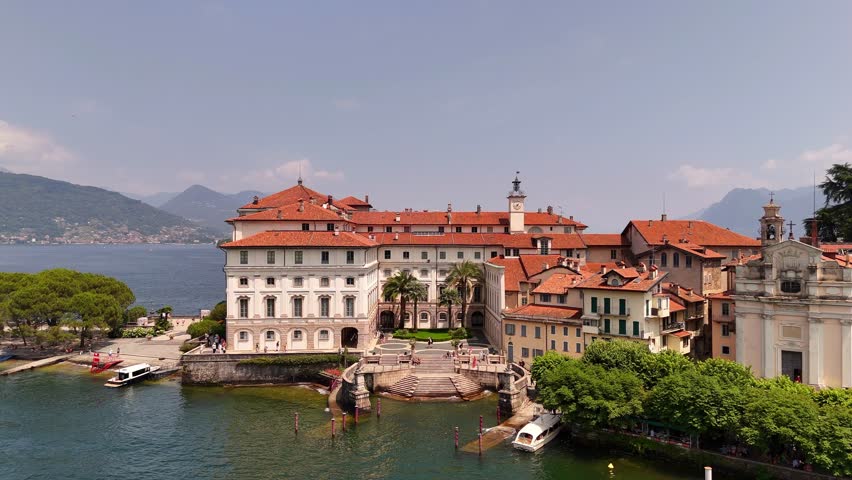Drone video of Isola Bella’s Borromeo Palace seen from the lake. Elegant red-roofed estate rises above the water, surrounded by Alpine peaks, lush gardens, and sparkling Lake Maggiore, Italy