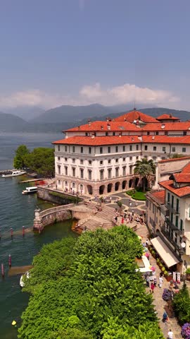 Drone video of Isola Bella’s Borromeo Palace seen from the lake. Elegant red-roofed estate rises above the water, surrounded by Alpine peaks, lush gardens, and sparkling Lake Maggiore, Italy