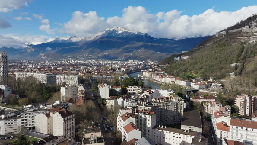 Aerial view of Grenoble cityscape revealing snow capped French Alps, Isere River, urban landscape with verdant spaces and architectural elements under bright sunlight