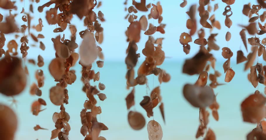 Artistic shot of boho-style seashell strands swaying against soft turquoise ocean, creating a calming and aesthetic ambiance for lifestyle, spa, or summer travel content.